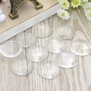 May include: Ten clear glass cabochons, also known as glass domes, are arranged on a light wood surface. The cabochons are round and have a smooth, polished finish.