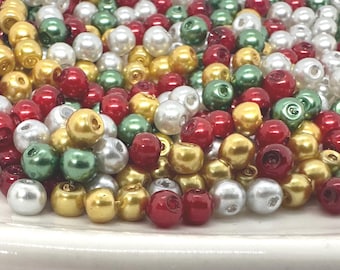 May include: A collection of colorful beads in various shades of red, gold, green, and white. The beads are round and have a glossy finish, suggesting they are made of glass or a similar material. They are scattered across a white surface.