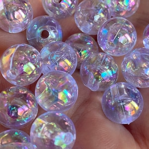 May include: A close-up of iridescent clear plastic beads. The beads are round and have a holographic finish. They are scattered on a hand.