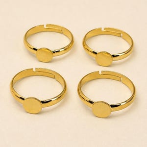 May include: Four gold-toned adjustable rings, each featuring a smooth, circular flat plate setting on the front. The bands are open at the back, allowing for size adjustment, and have a polished, reflective metallic finish throughout the design.