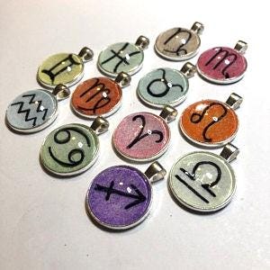 May include: A collection of twelve round silver-toned metal pendants, each featuring a unique black zodiac symbol set against a colorful, textured background. The pendants are arranged in a cluster, showcasing various colors including purple, orange, pink, blue, green, and yellow.