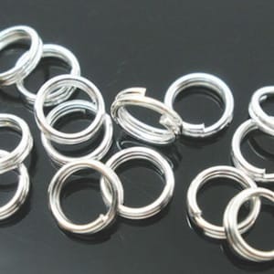 May include: A group of silver jump rings, a type of jewellery finding, are scattered on a black surface. The rings are all the same size and have a smooth, polished finish.