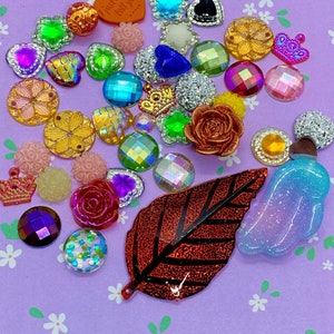 May include: A selection of craft embellishments in various shapes and colours, including rhinestones, heart-shaped gems, and flower-shaped beads. A red leaf and a heart-shaped charm with text are also present. The items are on a light purple surface.