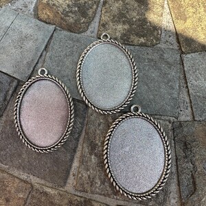 May include: Three oval pendant trays with silver-coloured rope-like edging. The inner surfaces have a glittery appearance in shades of pink and silver. Each tray has a small loop at the top for attaching a chain or cord.