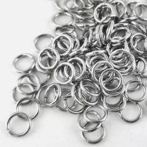 May include: A collection of small, silver-coloured jump rings. These metal rings are open and ready for use in jewellery making or other craft projects. The rings are scattered across a white surface.