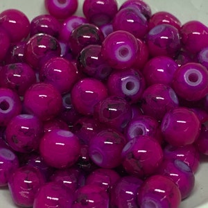 May include: A collection of vibrant, round beads in shades of magenta and deep purple. Each bead has a glossy finish and a unique pattern of darker swirls and a white center, suggesting a natural stone or glass material. These beads are ideal for jewelry making.