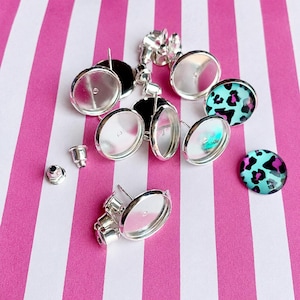 May include: A collection of silver-toned metal earring settings with circular bezels, some empty and some featuring a vibrant turquoise and magenta leopard print pattern. Several small metal earring backs are scattered among the components.