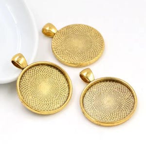 May include: Three circular gold-toned pendant trays featuring a textured, geometric pattern on the interior surface. Each pendant tray includes a small, integrated loop at the top for attachment, designed for jewelry making and crafting projects.