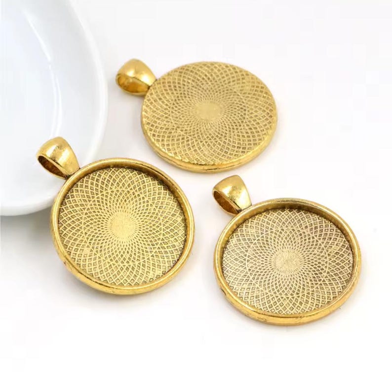 May include: Three circular gold-toned pendant trays featuring a textured, geometric pattern on the interior surface. Each pendant tray includes a small, integrated loop at the top for attachment, designed for jewelry making and crafting projects.