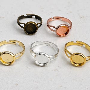 May include: Five adjustable metal rings with circular bezels for cabochon settings. The collection includes one antique bronze, one copper, one bright gold, one silver, and one yellow gold tone ring, each featuring a split band design.