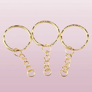 May include: Three gold keyrings with a circular ring and a chain attached. The keyrings are on a pink background.