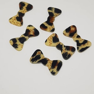 May include: Six small, brown, black, and yellow bow-shaped resin embellishments with a leopard print pattern.