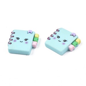 May include: Two light blue notebook charms with a smiling face, pink cheeks, and colourful accents. The charms have a spiral binding and a small bookmark.