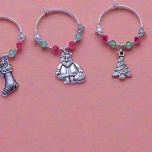 May include: Set of eight Christmas-themed wine glass charms. Each charm features a different holiday design, including a snowman, a stocking, a reindeer, a Christmas tree, a Santa Claus, a bell, an angel, and a sleigh. The charms are made of silver metal and are decorated with red and green beads.