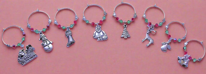 May include: Set of eight Christmas-themed wine glass charms. Each charm features a different holiday design, including a snowman, a stocking, a reindeer, a Christmas tree, a Santa Claus, a bell, an angel, and a sleigh. The charms are made of silver metal and are decorated with red and green beads.