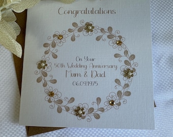 May include: A square white greeting card featuring a brown floral wreath design with embedded gold and pearl-like embellishments. The text reads: Congratulations, On Your 50th Wedding Anniversary, Mum & Dad, 06.09.1975. The card is placed on a brown envelope.