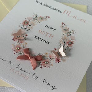 May include: A white birthday card with a floral wreath design, featuring the text "Happy 60th Birthday" and "To a Wonderful Mum". The card includes pink butterflies, a bow, and the date 12.10.2025.