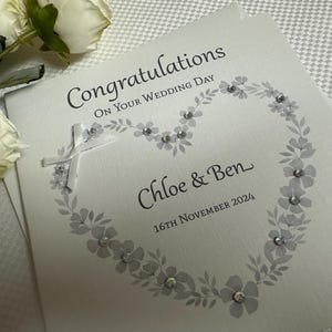 May include: A square white wedding greeting card featuring the text Congratulations On Your Wedding Day, Chloe & Ben, and 16th November 2024. The card is decorated with a grey floral heart illustration, small clear crystals, and a white ribbon cross.