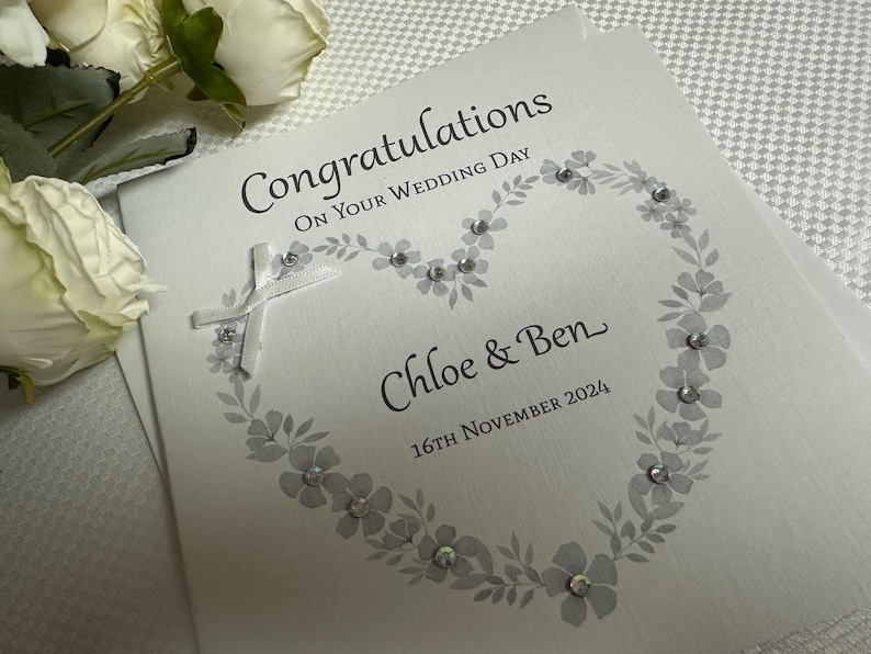 May include: A square white wedding greeting card featuring the text Congratulations On Your Wedding Day, Chloe & Ben, and 16th November 2024. The card is decorated with a grey floral heart illustration, small clear crystals, and a white ribbon cross.