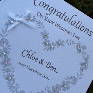 May include: A white wedding greeting card featuring the text "Congratulations ON YOUR WEDDING DAY", "Chloe & Ben", and "16TH NOVEMBER 2024". The card displays a gray floral heart design accented with iridescent rhinestones and a white ribbon bow.