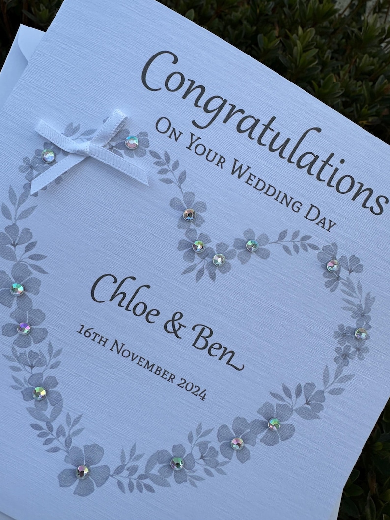 May include: A white wedding greeting card featuring the text "Congratulations ON YOUR WEDDING DAY", "Chloe & Ben", and "16TH NOVEMBER 2024". The card displays a gray floral heart design accented with iridescent rhinestones and a white ribbon bow.