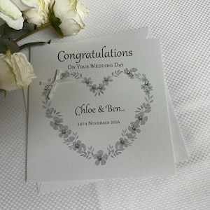 May include: A square white wedding greeting card featuring the text Congratulations On Your Wedding Day, Chloe & Ben, and 16th November 2024. The card displays a gray floral heart design accented with small clear crystals and a white ribbon bow.