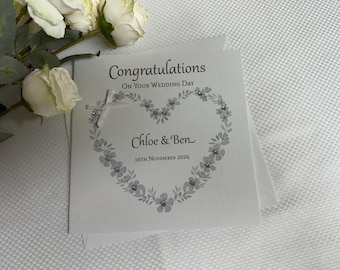 Puede incluir: Una tarjeta de felicitación de boda cuadrada de color blanco con el texto Congratulations On Your Wedding Day, Chloe & Ben, y 16th November 2024. La tarjeta muestra un diseño de corazón floral gris con pequeños cristales y un lazo blanco.