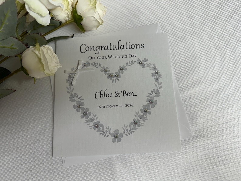 May include: A square white wedding greeting card featuring the text Congratulations On Your Wedding Day, Chloe & Ben, and 16th November 2024. The card displays a gray floral heart design accented with small clear crystals and a white ribbon bow.