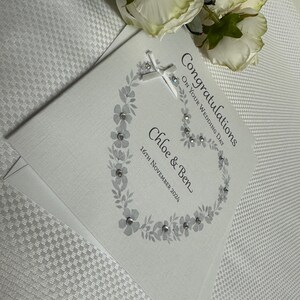 May include: A white square wedding card featuring a gray floral wreath design accented with small clear crystals. The text reads Congratulations ON YOUR WEDDING DAY, Chloe & Ben, and 16TH NOVEMBER 2024. A small white ribbon bow is attached at the top.