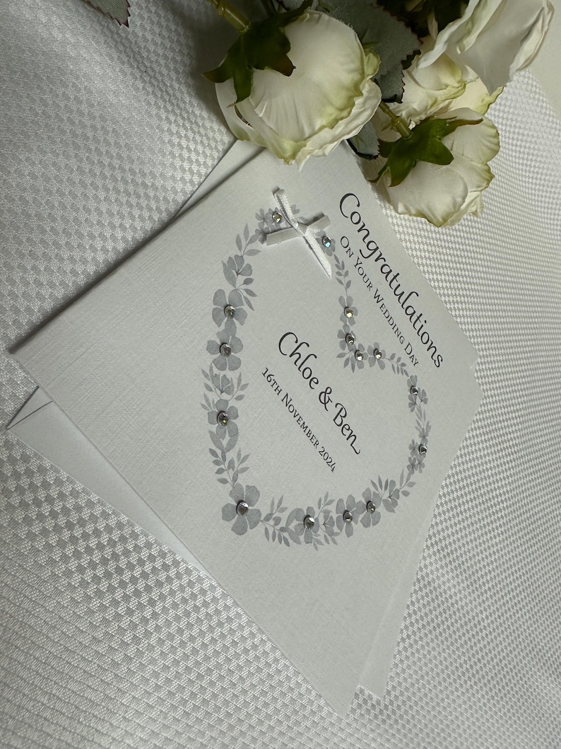 May include: A white square wedding card featuring a gray floral wreath design accented with small clear crystals. The text reads Congratulations ON YOUR WEDDING DAY, Chloe & Ben, and 16TH NOVEMBER 2024. A small white ribbon bow is attached at the top.
