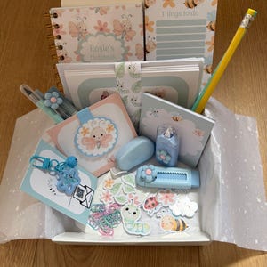 May include: A stationery gift set in a white box, featuring a spiral notebook, notepads, a pencil, pens, stickers, a keychain, and a blue eraser. The notebook and notepads have butterfly and bee designs. The box has a light blue interior.