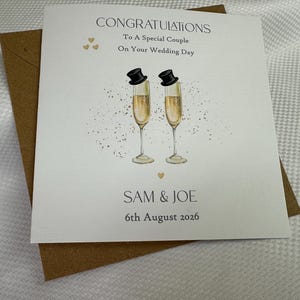 Pode incluir: Um cart&atilde;o de felicita&ccedil;&otilde;es de casamento quadrado branco com um envelope castanho. O cart&atilde;o apresenta duas flutes de champanhe com cartolas pretas, cora&ccedil;&otilde;es dourados e o texto: CONGRATULATIONS To A Special Couple On Your Wedding Day SAM & JOE 6th August 2026.