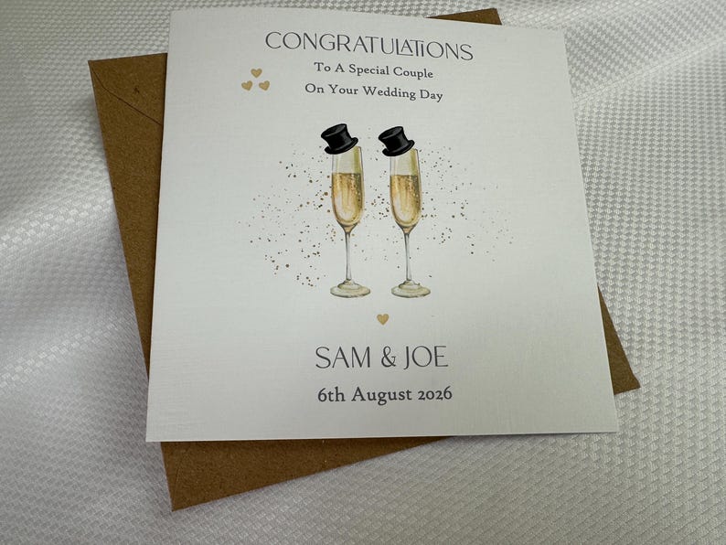 Pode incluir: Um cart&atilde;o de felicita&ccedil;&otilde;es de casamento quadrado branco com um envelope castanho. O cart&atilde;o apresenta duas flutes de champanhe com cartolas pretas, cora&ccedil;&otilde;es dourados e o texto: CONGRATULATIONS To A Special Couple On Your Wedding Day SAM & JOE 6th August 2026.