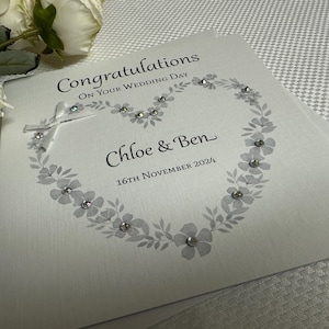 May include: A white wedding greeting card featuring the text Congratulations On Your Wedding Day, Chloe & Ben, and 16th November 2024. The card displays a printed gray floral heart design embellished with small clear crystals and a white ribbon bow.