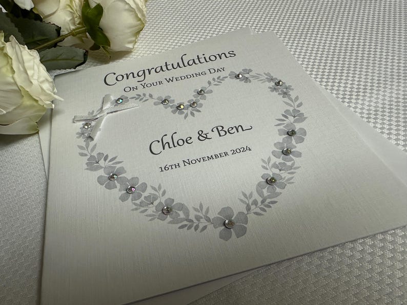 May include: A white wedding greeting card featuring the text Congratulations On Your Wedding Day, Chloe & Ben, and 16th November 2024. The card displays a printed gray floral heart design embellished with small clear crystals and a white ribbon bow.
