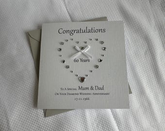 May include: A square white greeting card featuring the text Congratulations, 60 Years, To A Special Mum & Dad, On Your Diamond Wedding Anniversary, and 17.11.1966. The card is decorated with a heart shape made of small crystals and a white ribbon bow.