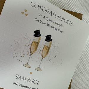 Pode incluir: Um cart&atilde;o de felicita&ccedil;&otilde;es de casamento quadrado em cartolina branca com o texto CONGRATULATIONS To A Special Couple On Your Wedding Day, SAM & JOE e 6th August 2026. O design inclui duas ta&ccedil;as de champanhe com cartolas pretas e cora&ccedil;&otilde;es dourados.