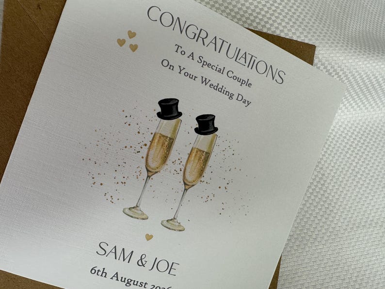 Pode incluir: Um cart&atilde;o de felicita&ccedil;&otilde;es de casamento quadrado em cartolina branca com o texto CONGRATULATIONS To A Special Couple On Your Wedding Day, SAM & JOE e 6th August 2026. O design inclui duas ta&ccedil;as de champanhe com cartolas pretas e cora&ccedil;&otilde;es dourados.