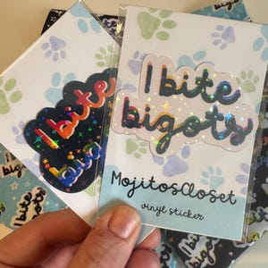 May include: A clear vinyl sticker with the text "I bite bigots" in a cloud-shaped design. The sticker has a holographic effect, with a light blue background and the text "MojitosCloset vinyl sticker".