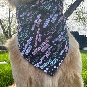 May include: Black dog bandana with the phrase "little bit of an ASSHOLE" in a repeating pattern. The text is in a mix of colors including pink, blue, and white. The bandana is triangular and worn around the dog's neck.