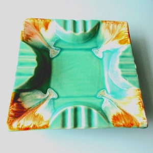 May include: A square ceramic ashtray featuring a glossy glaze in shades of turquoise, mint green, and dark green. The corners are accented with blended streaks of orange, yellow, and white, and the edges include four integrated cigarette rests.