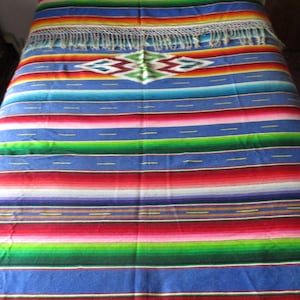 May include: A woven blanket featuring horizontal stripes in vibrant shades of blue, red, green, yellow, and white. The textile includes a central diamond-shaped pattern with green and maroon accents and is finished with long white fringe at the ends.