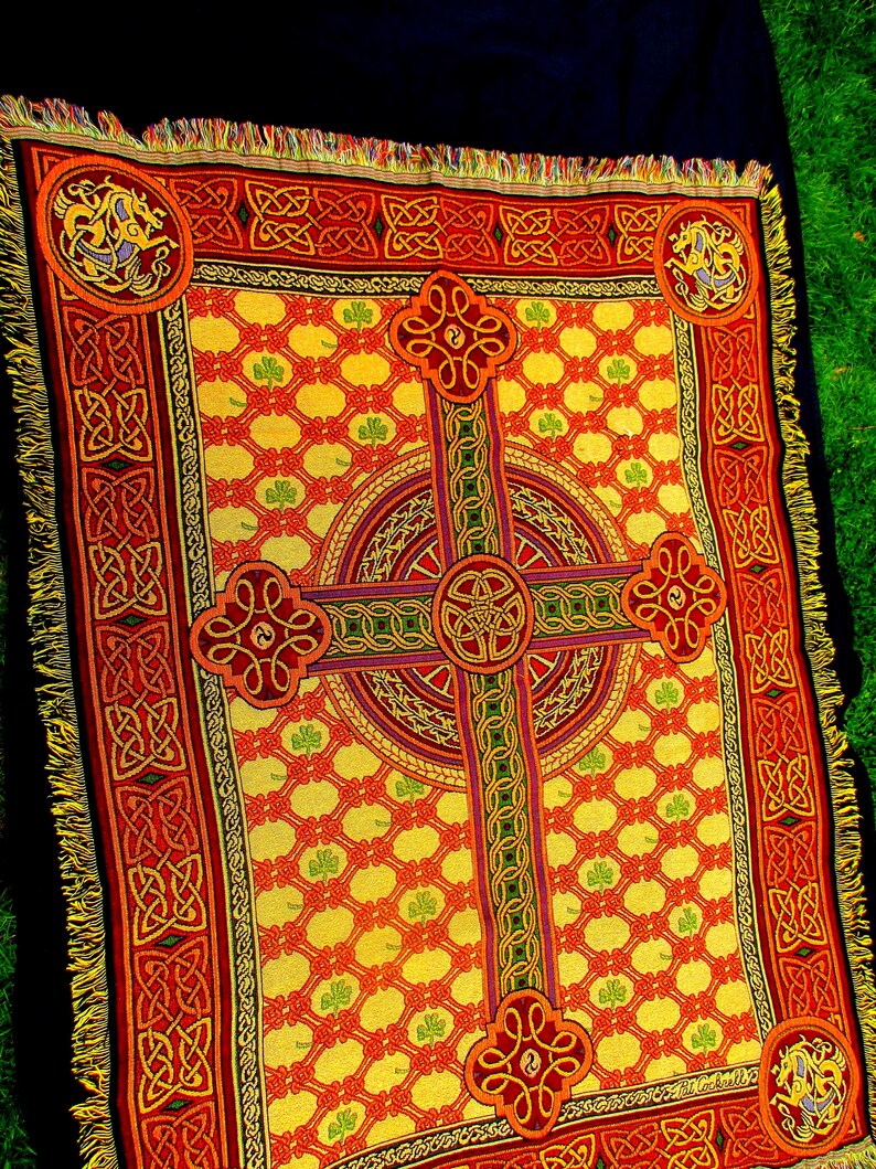 May include: A woven throw blanket featuring a prominent Celtic cross design in red, orange, and yellow. The background displays a repeating grid pattern with small green shamrocks. The border includes intricate knotwork and corner medallions.