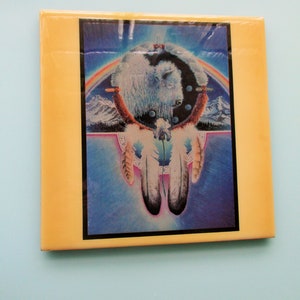 May include: A yellow square tile with a colorful image of a white buffalo inside a dreamcatcher with feathers. The dreamcatcher has a rainbow arch above it and a blue background.