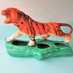 May include: A ceramic tiger figurine with orange fur and black stripes, featuring an open mouth and a metal chain collar. The tiger stands atop a green, textured log-shaped base that contains three hollow openings for holding small items.