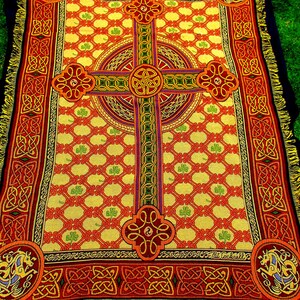 May include: A rectangular woven throw blanket featuring a prominent central Celtic cross design in red, orange, and green on a yellow background. The pattern includes repeating shamrock motifs, intricate knotwork borders, and corner medallions.