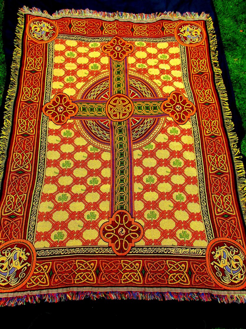 May include: A rectangular woven throw blanket featuring a prominent central Celtic cross design in red, orange, and green on a yellow background. The pattern includes repeating shamrock motifs, intricate knotwork borders, and corner medallions.
