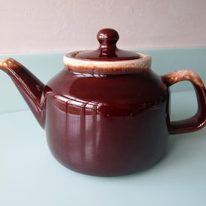 May include: A glossy dark brown teapot with a matching lid. The teapot features a rounded body, a curved spout, and a sturdy handle. The rim of the lid and the top edges of the spout and handle are accented with a lighter, creamy beige drip glaze.