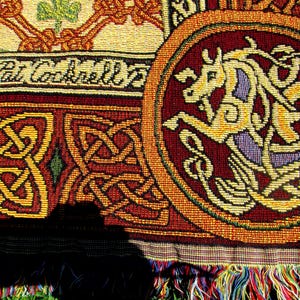 May include: A woven textile featuring a circular medallion with a stylized horse in yellow and purple against a deep red background. The fabric displays intricate Celtic knotwork patterns in red and gold, with the text Pat Cockrell visible.