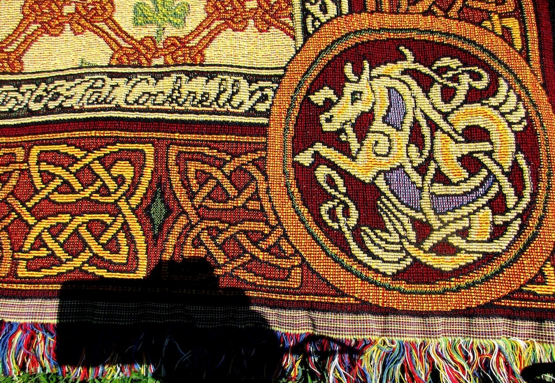 May include: A woven textile featuring a circular medallion with a stylized horse in yellow and purple against a deep red background. The fabric displays intricate Celtic knotwork patterns in red and gold, with the text Pat Cockrell visible.